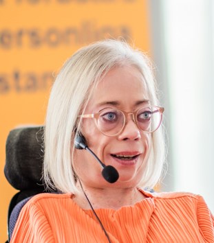 Sophie Karlssons ansiktes syns, hon har en mikrofon längs med kinden,glasögon, pratar. Oranga tröja