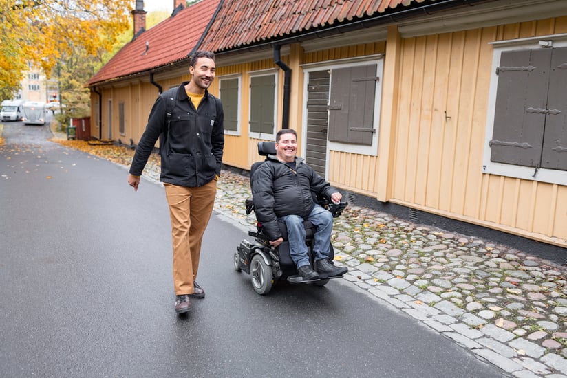 Bild på en assistansanvändare och en personlig assistent på promenad. Båda är glada. 