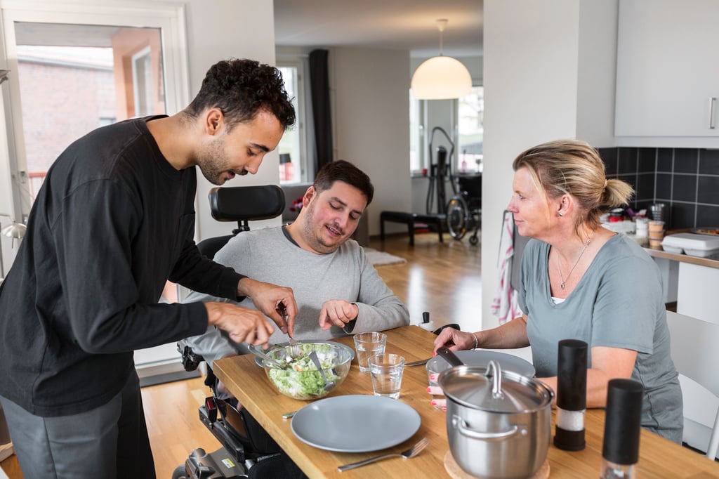 En assistent assisterar en man med att kära upp maten, de sitter vid köksbordet. Bredvid sitter en kvinna och pratar med mannen.