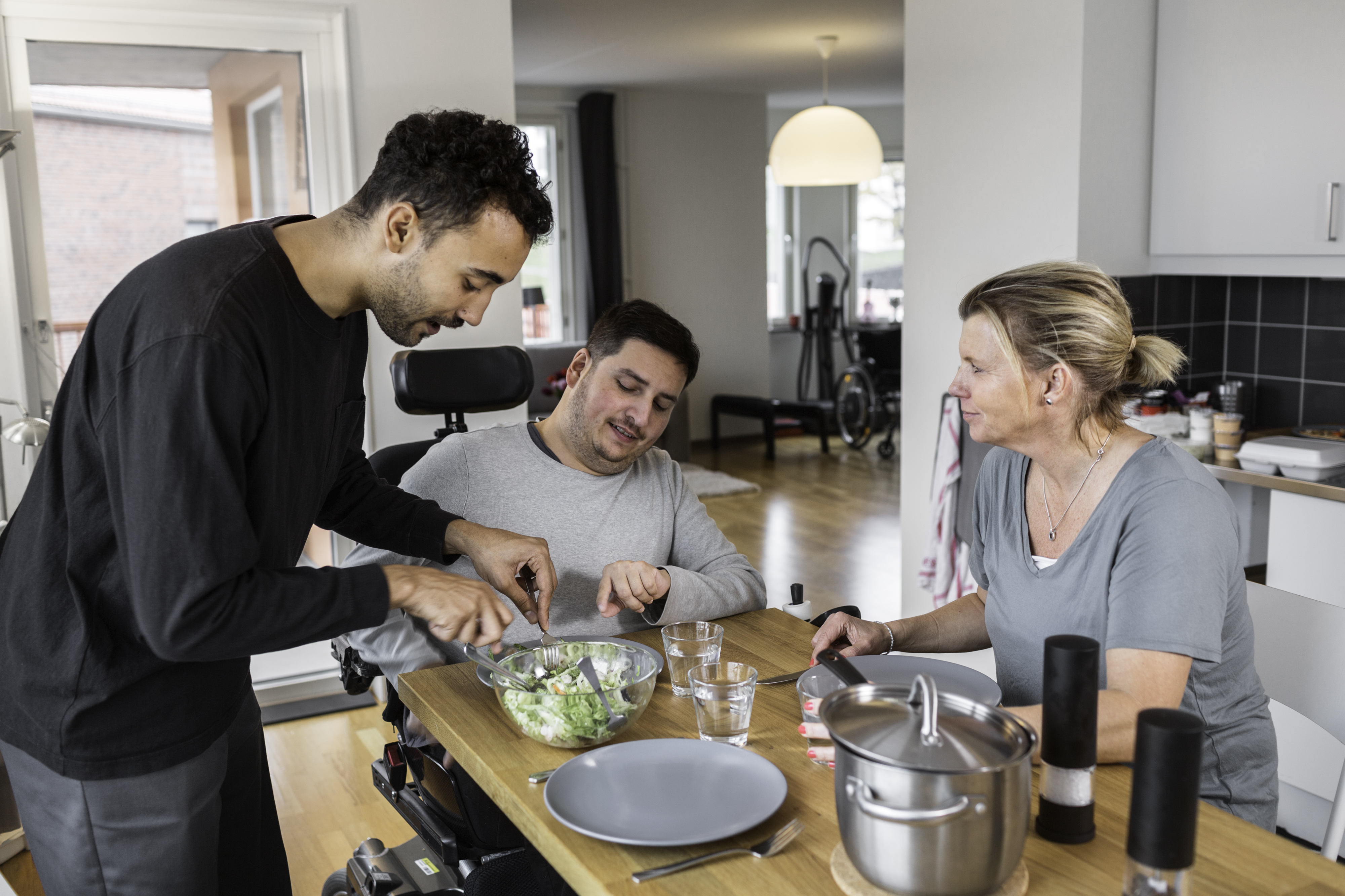 En assistent assisterar en man med att kära upp maten, de sitter vid köksbordet. Bredvid sitter en kvinna och pratar med mannen.